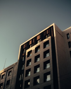 A modern multi-story building with a geometric facade featuring square windows. The exterior is composed of light-colored materials, contrasting with vertical dark elements. The lighting creates shadows and emphasizes the building's contemporary architectural design.
