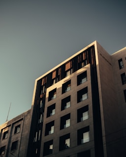 A modern multi-story building with a geometric facade featuring square windows. The exterior is composed of light-colored materials, contrasting with vertical dark elements. The lighting creates shadows and emphasizes the building's contemporary architectural design.