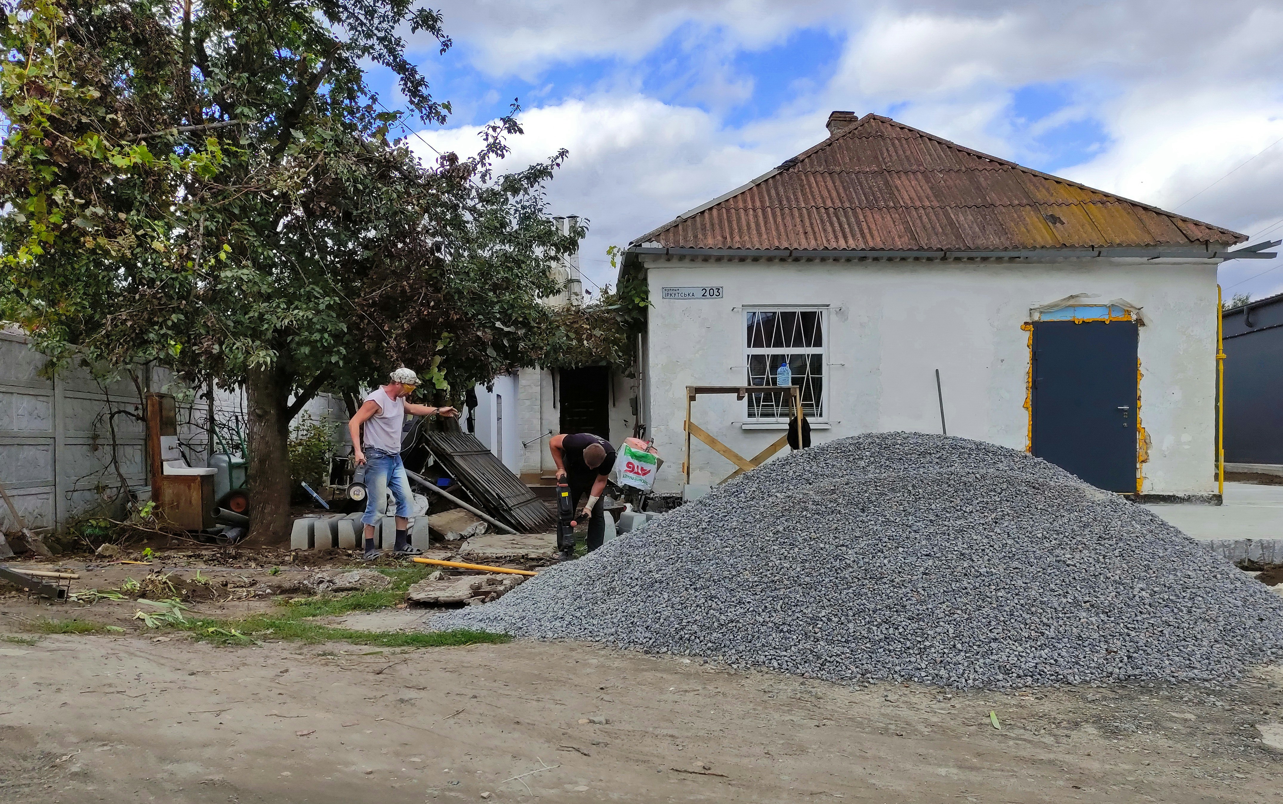 A small, old house with a rusted roof and white walls stands with a pile of gravel in front. Two men are working near the gravel, one holding a tool, and surrounded by scattered building materials. A large tree with green leaves is on the left side.