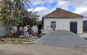 A small, old house with a rusted roof and white walls stands with a pile of gravel in front. Two men are working near the gravel, one holding a tool, and surrounded by scattered building materials. A large tree with green leaves is on the left side.