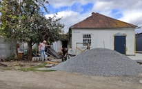 A small, old house with a rusted roof and white walls stands with a pile of gravel in front. Two men are working near the gravel, one holding a tool, and surrounded by scattered building materials. A large tree with green leaves is on the left side.