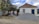 A small, old house with a rusted roof and white walls stands with a pile of gravel in front. Two men are working near the gravel, one holding a tool, and surrounded by scattered building materials. A large tree with green leaves is on the left side.