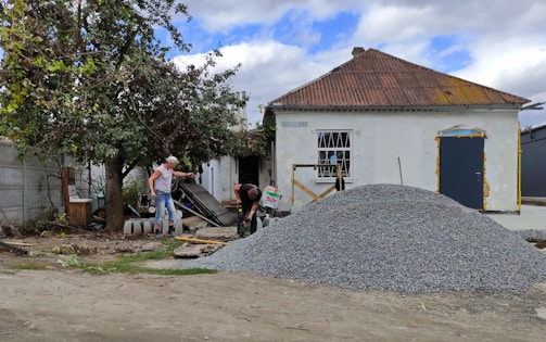 A small, old house with a rusted roof and white walls stands with a pile of gravel in front. Two men are working near the gravel, one holding a tool, and surrounded by scattered building materials. A large tree with green leaves is on the left side.