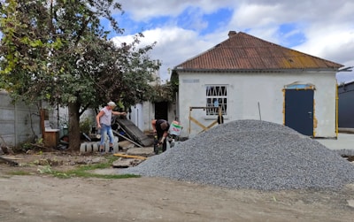 A small, old house with a rusted roof and white walls stands with a pile of gravel in front. Two men are working near the gravel, one holding a tool, and surrounded by scattered building materials. A large tree with green leaves is on the left side.