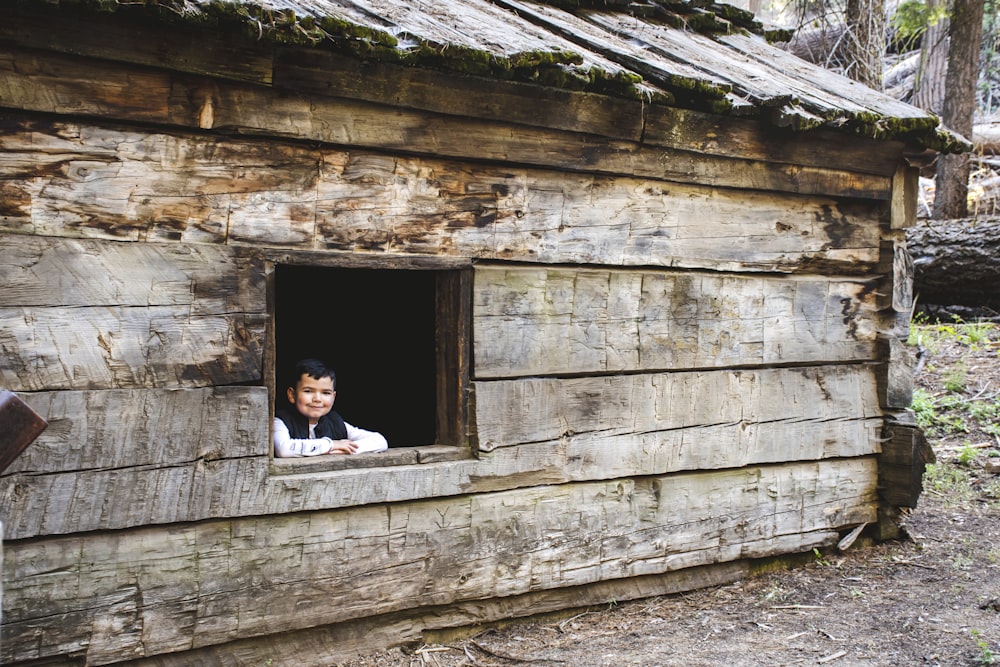 a young boy looking out of a window of a log cabin