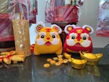 Two plush toy tigers, one yellow and the other red, sit on a dark surface surrounded by decorative gold ingots and coins. On the left, there is a detailed yellow cut-out cylindrical decoration and rolled decorative items wrapped in gold. In the background, wrapped gift baskets can be seen.