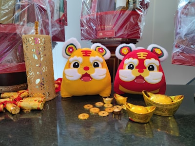 Two plush toy tigers, one yellow and the other red, sit on a dark surface surrounded by decorative gold ingots and coins. On the left, there is a detailed yellow cut-out cylindrical decoration and rolled decorative items wrapped in gold. In the background, wrapped gift baskets can be seen.