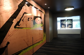 The image features an indoor corridor with contemporary art pieces on the walls. On the left, a large mural with abstract designs and geometric patterns in shades of orange, black, and green. At the end of the corridor, a painting or print of intense eyes displayed on a window or glass panel adds a dramatic effect.