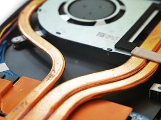 Close-up view of the internal components of an electronic device, featuring copper heat pipes, a cooling fan, and circuit board parts. The image highlights the intricate design and arrangement of these components.