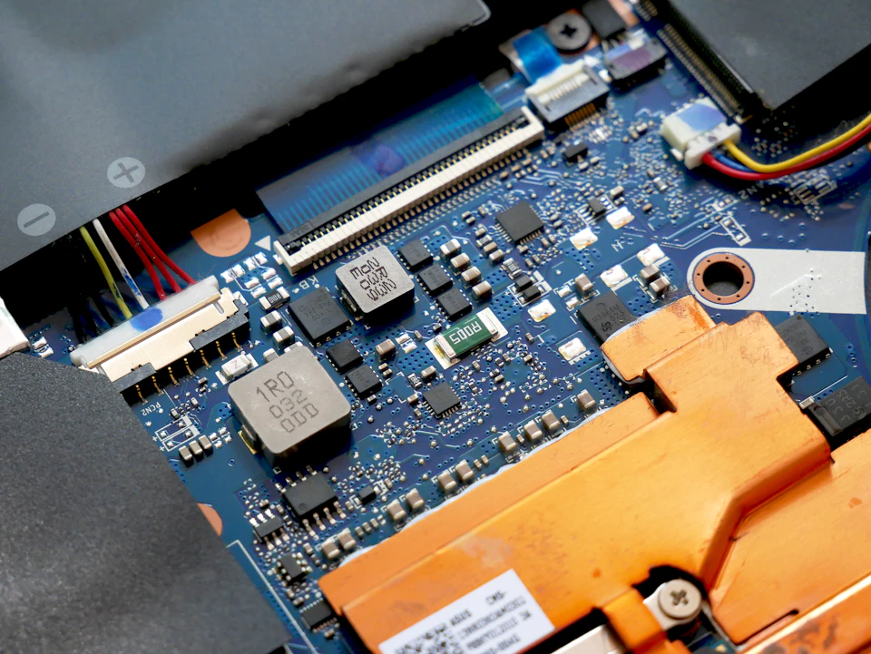 The image shows a close-up view of an internal computer circuit board, specifically focusing on the blue and white components. The circuit board is black with various colored wires attached to it, indicating its complex nature. The main component visible in the image is a small chip, possibly a microchip or a component used for data processing. The background of the image is blurred, drawing attention to the intricate details of the circuit board.