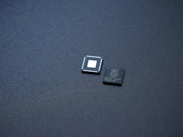 Two small integrated circuits are placed on a textured, dark surface. One of the chips has visible pins along its edges and a blank square in the center, while the other has markings and logos printed on it.