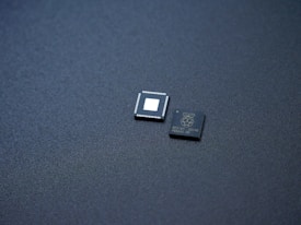 Two small integrated circuits are placed on a textured, dark surface. One of the chips has visible pins along its edges and a blank square in the center, while the other has markings and logos printed on it.