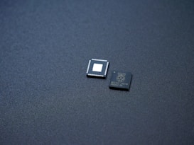 Two small integrated circuits are placed on a textured, dark surface. One of the chips has visible pins along its edges and a blank square in the center, while the other has markings and logos printed on it.