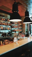 A cozy bar interior features a well-organized selection of liquor bottles on shelves behind the counter, illuminated by hanging pendant lights. The counter displays various bar tools, a small flower vase, and a QR code card. The ambiance is warm and inviting, with a stylish tiled wall in the background.