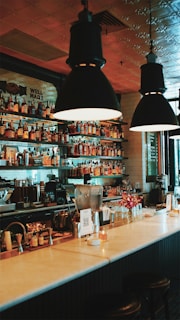 A cozy bar interior features a well-organized selection of liquor bottles on shelves behind the counter, illuminated by hanging pendant lights. The counter displays various bar tools, a small flower vase, and a QR code card. The ambiance is warm and inviting, with a stylish tiled wall in the background.
