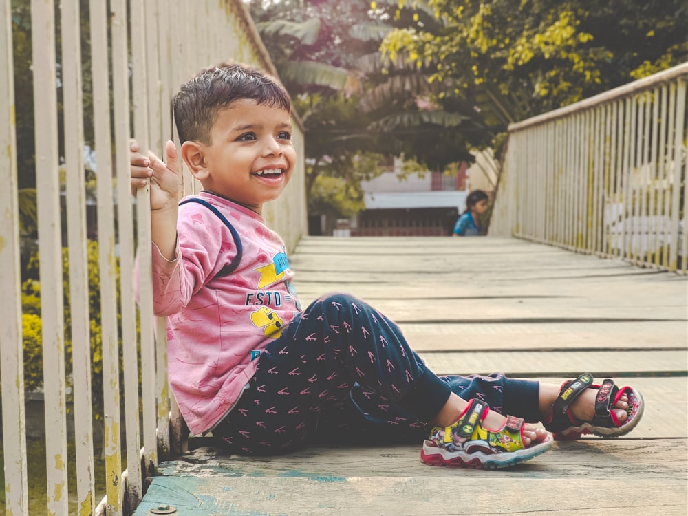 a little boy sitting on the ground next to a fence