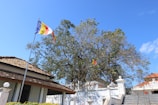A Buddhist flag flutters on a pole in front of a tiled-roof building. Behind the building, there are large trees with green leaves under a clear blue sky. Stone steps lead up to a structure with white walls and a decorative fence.