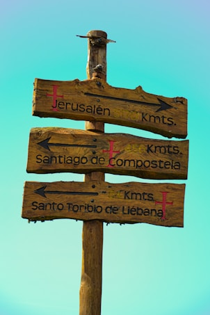 A wooden signpost with three directional arrows carved into the wood, indicating distances to different locations: Jerusal&eacute;n, Santiago de Compostela, and Santo Toribio de Li&eacute;bana. The wood appears aged and weathered, and each sign has a red cross emblem.