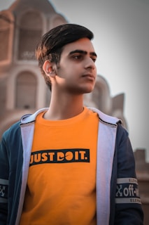 A young person with short dark hair is wearing an orange shirt with bold lettering and a denim-like jacket. The background features an architectural structure with arches, giving a historic or cultural context.