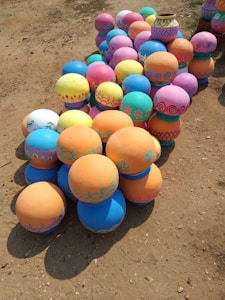 A collection of brightly colored and intricately painted clay pots arranged on a sandy ground. The pots feature a variety of patterns, including floral designs and geometric shapes, utilizing vivid shades of blue, pink, yellow, orange, and teal.