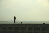 A photographer stands on a concrete pier with a camera mounted on a tripod, facing a distant skyline across the water under a hazy sky.