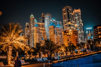 A vibrant cityscape at night featuring illuminated skyscrapers, palm trees, and a pool area with lounge chairs. The buildings are adorned with lights, creating a lively and luxurious atmosphere.