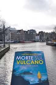 A book cover titled 'Morte Sul Vulcano' is held up in the foreground, featuring a person wearing a yellow coat standing in front of a volcano. In the background, there is a canal lined with traditional European buildings under a cloudy sky.