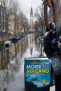 A picturesque canal scene with boats docked along the sides, flanked by tall, narrow buildings typical of European architecture. In the distance, a towering church or clock tower rises against a cloudy sky. In the foreground, a book titled 'Morte Sul Vulcano' with a mountainous cover is placed prominently, adding a thematic element to the serene surroundings.