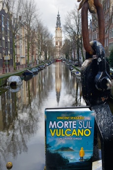 A picturesque canal scene with boats docked along the sides, flanked by tall, narrow buildings typical of European architecture. In the distance, a towering church or clock tower rises against a cloudy sky. In the foreground, a book titled 'Morte Sul Vulcano' with a mountainous cover is placed prominently, adding a thematic element to the serene surroundings.