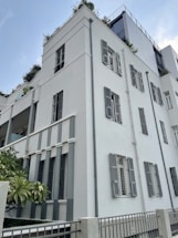 A modern multi-story building with clean, geometric lines and light-colored walls. The structure features numerous windows with shutters and some balcony areas adorned with plants. The architecture blends contemporary design with natural elements, creating a balanced and aesthetically pleasing appearance.