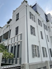 A modern multi-story building with clean, geometric lines and light-colored walls. The structure features numerous windows with shutters and some balcony areas adorned with plants. The architecture blends contemporary design with natural elements, creating a balanced and aesthetically pleasing appearance.