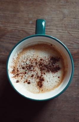 A teal mug filled with frothy coffee, topped with sprinkles of cocoa or cinnamon. The mug is placed on a textured wooden surface, creating a cozy and inviting atmosphere.