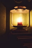A vintage-style lantern with a glass enclosure contains a lit candle that emits a warm, soft glow. The lantern is placed against a dark backdrop, creating an intimate and cozy atmosphere with gentle shadows.