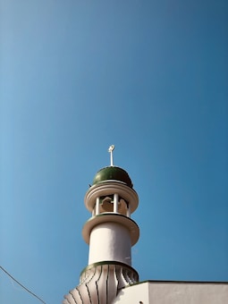 A mosque tower with a green dome and crescent moon finial against a clear blue sky. The structure features white and green accents with architectural details typical of Islamic design.