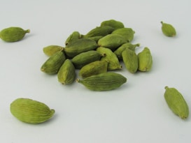 A collection of green cardamom pods is scattered across a light surface. The pods have a ribbed texture and an elongated shape, with some clustered closely together and others spread out.