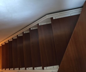 A wooden staircase viewed from a steep angle, with each step having a metal trim on the edges. The lighting is dim, and the staircase appears to be in an enclosed space with a low ceiling.