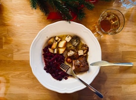 A white plate on a wooden table contains a meal with roasted potatoes, red cabbage, and what appears to be roasted meat and vegetables. A glass of amber liquid stands nearby, and some green fir branches are visible at the top.