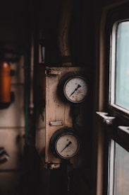 A pair of old, round pressure gauges mounted on a wall. The gauges have an industrial and slightly weathered appearance, with one needle pointing at a specific measurement. A window on the right allows dim light to enter, casting shadows and adding depth to the scene.