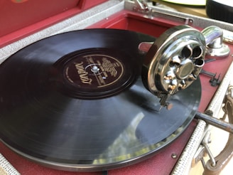 A close-up view of an old-fashioned gramophone playing a black vinyl record. The record player features a detailed metal tonearm and needle, with a red and beige textured casing.