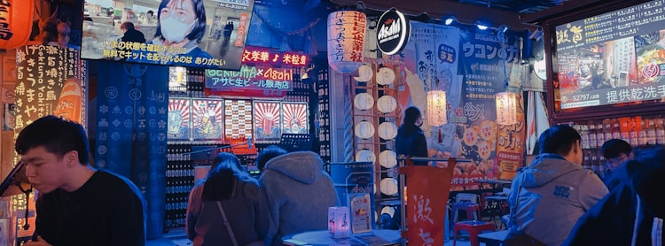 The interior of a vibrant Japanese-themed restaurant or bar is filled with colorful neon signs and posters in Japanese script. People are seated at tables, with a few visible screens displaying broadcasts. The atmosphere is lively with an array of lanterns and ornate decorations creating an immersive ambiance.