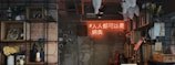 A cozy, rustic interior featuring a wall with exposed brick and concrete surfaces, adorned with wooden shelves displaying various decorative items like vases, candles, potted plants, and framed pictures. A neon sign with text in an Asian script glows in warm orange hues, adding a modern touch. There's a red metal chair and various knick-knacks that suggest a creative and eclectic environment.