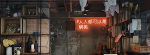 A cozy, rustic interior featuring a wall with exposed brick and concrete surfaces, adorned with wooden shelves displaying various decorative items like vases, candles, potted plants, and framed pictures. A neon sign with text in an Asian script glows in warm orange hues, adding a modern touch. There's a red metal chair and various knick-knacks that suggest a creative and eclectic environment.