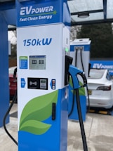 A modern electric vehicle charging station is prominently displayed with the branding 'EV Power' and a capacity of 150kW. The charger features a digital screen and contactless payment options. In the background, an electric car is partially visible under a canopy.