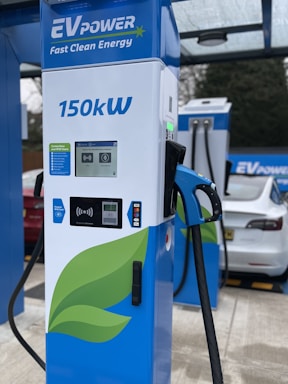 A modern electric vehicle charging station is prominently displayed with the branding 'EV Power' and a capacity of 150kW. The charger features a digital screen and contactless payment options. In the background, an electric car is partially visible under a canopy.