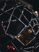 A close-up view of an intricately woven metallic structure in a geodesic pattern, with a complex mesh of interconnected segments. Light filters through the gaps, casting a warm, dim glow against a dark background, creating a dramatic contrast.