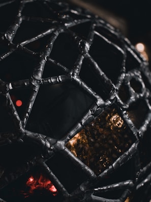 A close-up view of an intricately woven metallic structure in a geodesic pattern, with a complex mesh of interconnected segments. Light filters through the gaps, casting a warm, dim glow against a dark background, creating a dramatic contrast.