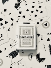 A deck of Navigators playing cards is placed at the center of a scattered arrangement of various playing cards. The cards feature different suits including hearts, spades, diamonds, and clubs. Some of the number and face cards are visible, with a queen in the upper left quadrant and jokers visible as well.
