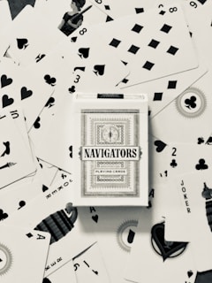 A deck of Navigators playing cards is placed at the center of a scattered arrangement of various playing cards. The cards feature different suits including hearts, spades, diamonds, and clubs. Some of the number and face cards are visible, with a queen in the upper left quadrant and jokers visible as well.