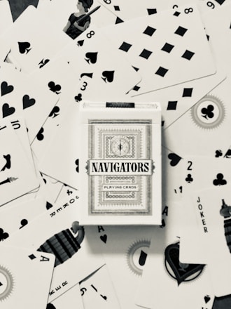 A deck of Navigators playing cards is placed at the center of a scattered arrangement of various playing cards. The cards feature different suits including hearts, spades, diamonds, and clubs. Some of the number and face cards are visible, with a queen in the upper left quadrant and jokers visible as well.