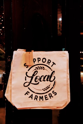 A canvas tote bag hangs against a dark background. The bag features bold, black text that reads 'Support Local Farmers,' accompanied by minimalist decorative elements like wheat stalks. There's a small tag attached to the bag's handle.